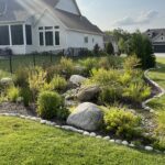 Backyard with garden, large rocks, shrubs, a gray house, metal fence, green grass, partly cloudy sky.