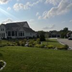 Residential area with a house, manicured lawn, garden, clear sky, clouds, and quiet street.